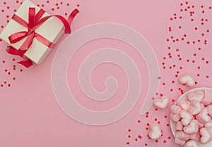 White gift box with red ribbon and heart-shaped marshmallows in white bowl on pink background with lots of small red hearts.
