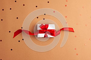 White gift box with a red satin ribbon bow tied on brown background with red stars shapes.Christmas Holidays and present concept