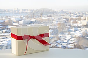 White gift box tied with a red ribbon on the background of the city. Horizontal composition with copy space. Great for Christmas