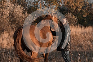 A white girl in leather jacket next to a horse and poses in the forest at sunset, like in a fairy tale. Well-groomed