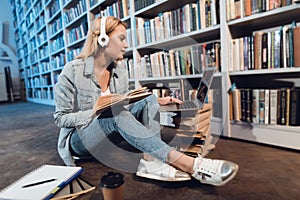 White girl near bookshelf in library. Student is listening to music, using laptop and reading book.