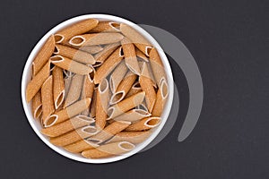 White glass bowl of raw brown penne rigate pasta. Isolated on a dark grey background.