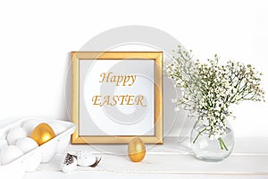 White and gold Easter eggs, a mockup with a gold frame and spring flowers in a vase on a white wooden table