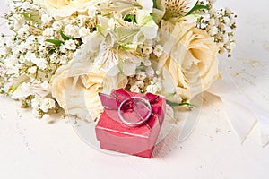 White gold ring rests on red gift box, bouquet of flowers on white textured wood background, holiday