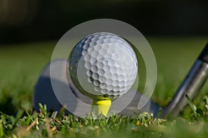 White golf ball teed up on a yellow tee with club face behind it and with soft green background