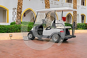 A white Golf cart is parked near the hotel complex. The electric car is charged in the Parking lot of the resort city. A car