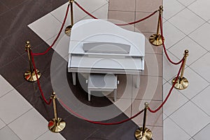 A white grand piano on a ceramic tiled floor is fenced with golden poles and red ropes. View from above