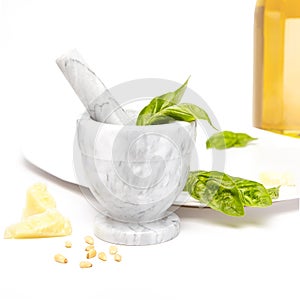 White and gray marble mortar and pestle with pesto ingredients, olive oil, basil, pine nuts, and parmesan cheese.