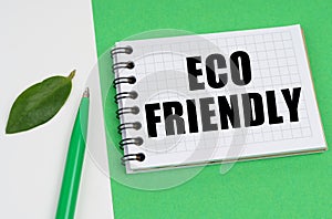 On a white-green background lies a pen, a leaf of a plant and a notepad with the inscription - ECO FRIENDLY