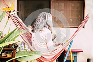 White hair diversity senior woman relax on an hammock in the garden at home reading a paper book and enjoying the quaity of