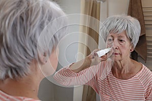 A white haired senior woman in her sixties is using a nasal spray in front of the bathroom mirror