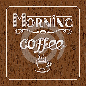 White Hand drawn lettering `Morning coffee` with charcoal effect and view of a cup of coffee on brown wood background.