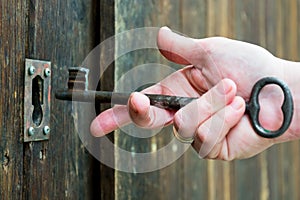 white hand putting an old or vintage key made by metallic iron inside a lock of a wooden door. The old key and the lock are