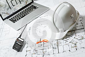 White hard hat, laptop  with drawings, glasses and walkie talkie with  and blueprints on a table