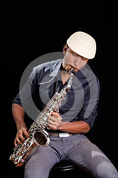 With white hat a black man is sitting and playing his saxophone, dark background, nice music