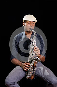 With white hat a black man is sitting and playing his saxophone, dark background, nice music