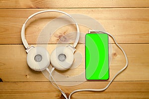 White headphones connected to phone on wooden table. Modern headphone with cable and gadget with green screen