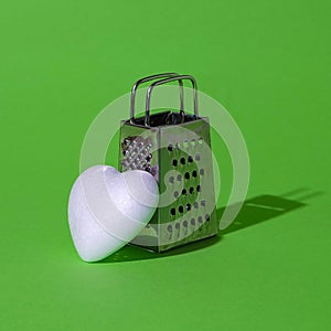 White heart and small grater on a green background. Harsh shadow. Love, life, brokenhearted, message, culinary arts, concept.