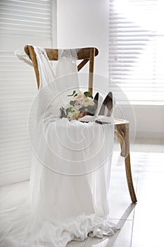 White high heel shoes, flowers and wedding dress on wooden chair indoors