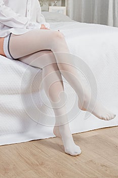 White hosiery. Beautiful long female legs in stockings. Girl putting on stockings at home in a white room. Varicose