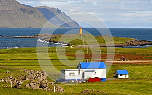A white house with a blue roof on the west coast of Iceland with an orange lighthouse and the sea and reef in the background.