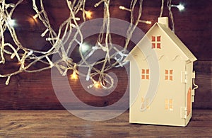 White house lantern with burning candles inside in front of garland gold lights on wooden table. retro filtered image