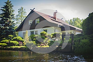 White house with a triangular roof on the river bank, surrounded by a garden with green plants and a well.