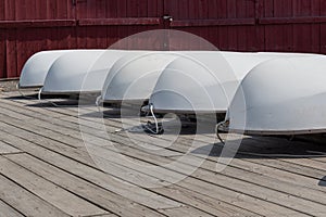 White hulls of small sailboats upside down on a wooden dock, red boathouse background