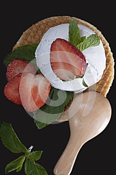 White ice cream with strawberries and mint on a waffle with a wooden spoon on a black background