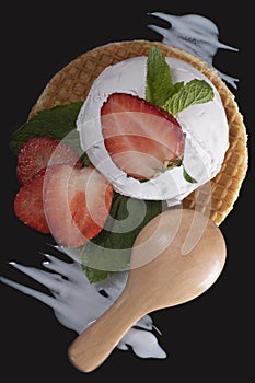 White ice cream with strawberries and mint on a waffle with a wooden spoon on a black surface with white cream