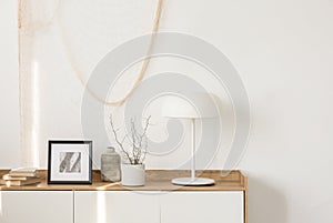 White industrial lamp, photo in frame and plant in pot on wooden console table in elegant living room with white wall