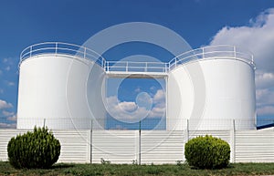White industrial liquid storage tanks with a white fence in front. Blue sky with fluffy clouds on background