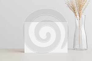 White invitation card mockup with a dried grass on a beige table. 5x7 ratio, similar to A6, A5