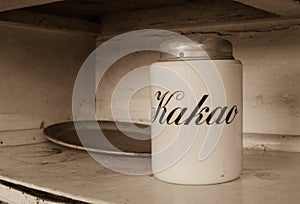 A white jar for storage  cocoa with text `Kakao` in russian languige and an old metal tray with rust. Old white kitchen shelf. Sep photo