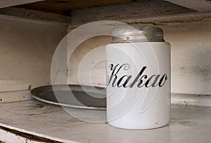 A white jar for storage  cocoa with text `Kakao` in russian languige and an old metal tray with rust. Old white kitchen shelf. Age photo