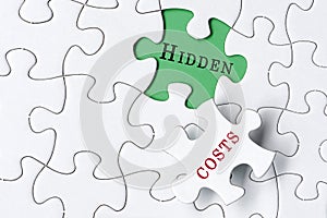 White jigsaw puzzle with word hidden costs over green background.