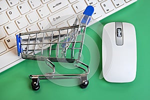 White keyboard and mouse for a computer on a green background. Shopping trolley lies on the keys. Concept online shopping, black