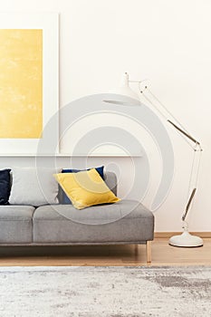 White lamp next to grey couch in modern living room interior with yellow poster and carpet. Real photo