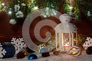 White lantern with a burning candle stands in the snow surrounded by Christmas decorations on the background of a wooden