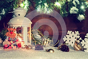 White lantern with a burning candle stands in the snow surrounded by Christmas decorations on the background of a wooden