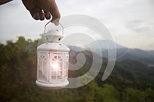 White lantern in hand on a mountains background. Morning view. Person carry lantern with candle light inside on mountain.