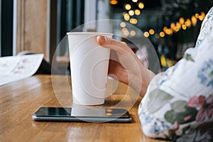 White large paper cup of tea on a table in a cafe. A woman`s hand holds the cup, a smartphone lies next to it.