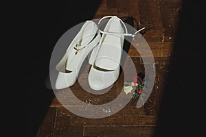 White leather high heels wedding shoes stand on the dark wooden floor. Small jewellery bracelet and earrings with buttonhole as