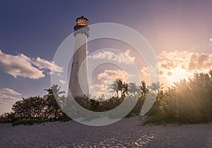 A white lighthouse against deep blue sky with setting sun tinting white clouds with orange pink light rays with white