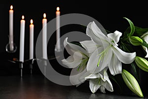 White lilies and blurred burning candles on table in darkness, closeup with space for text.