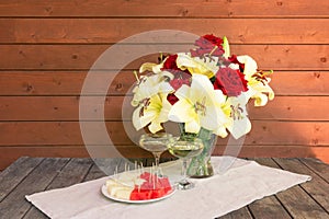 White lilies and red roses, glasses of champagne and plate with watermelon and melon on rustic wooden table