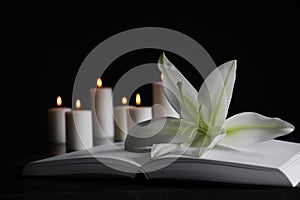 White lily, book and blurred burning candles on table in darkness, space for text. Funeral symbol