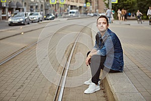 White male awaiting tram in the center of a European city, sitting on the sidewalk in dangerous proximity to the tracks