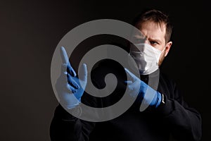 A white man with a beard in a medical mask and rubber gloves in black clothing points with his hands