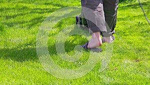White man in plastic slippers moving green lawn with cheap plastic electrical lawn mover at sunny day, closeup